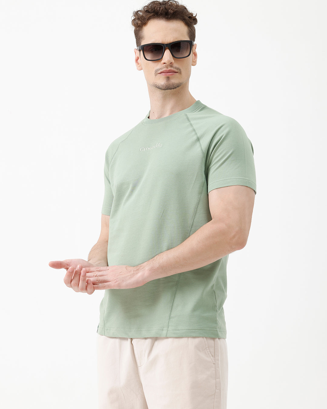 A man wearing a light sage green athletic t-shirt with a small tonal logo on the chest, paired with beige pants, standing against a plain white studio background.