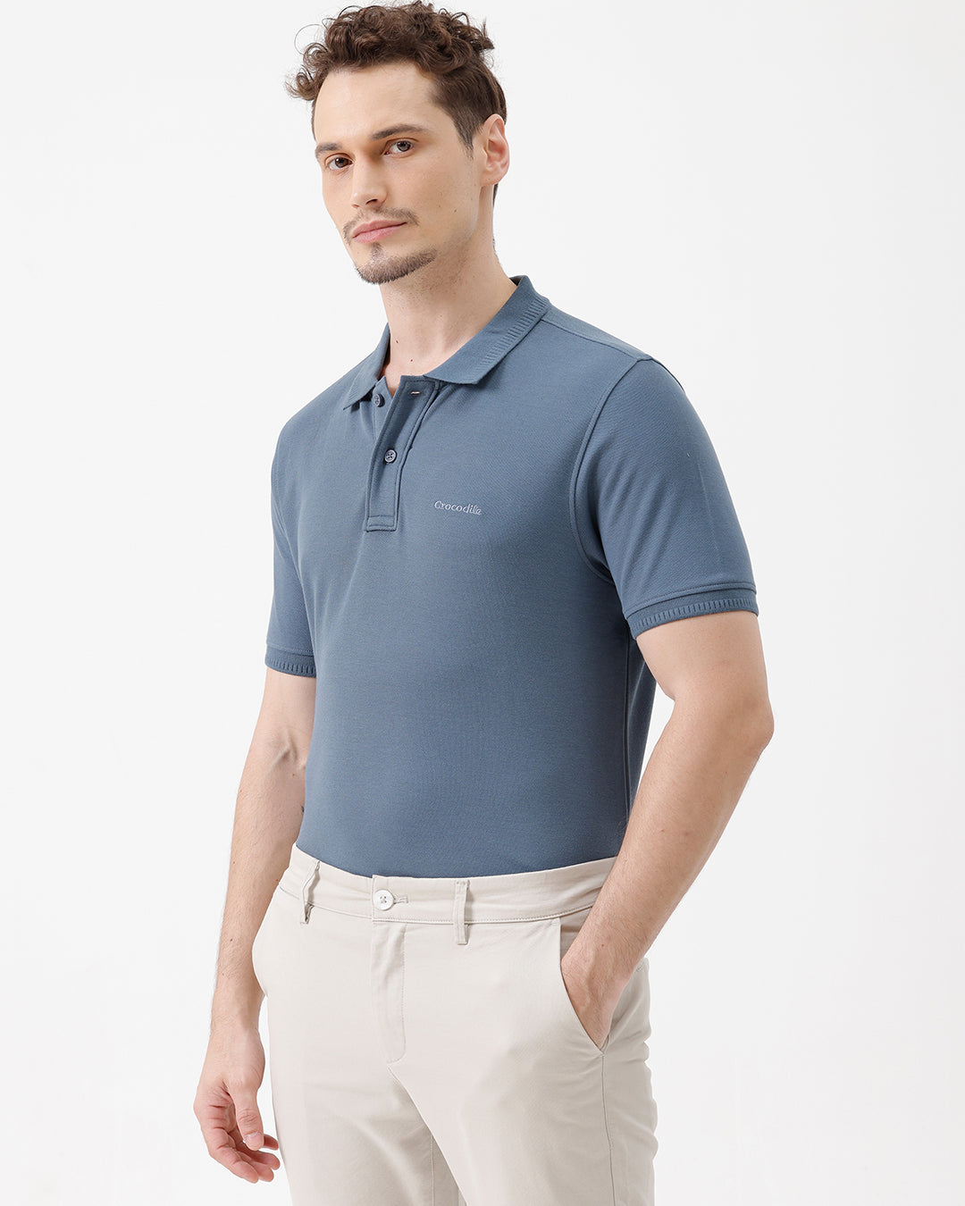 A man wearing a slate blue short-sleeve polo shirt and beige trousers, standing against a plain white studio background.