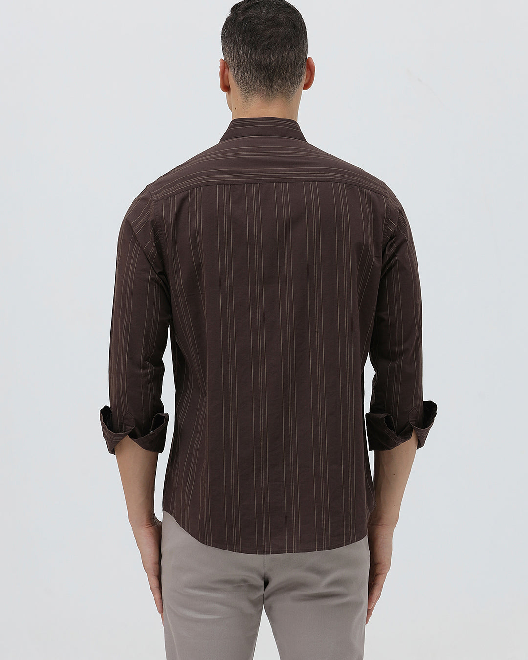 A man wearing a dark brown long-sleeve button-up shirt with thin vertical stripes and light gray trousers, standing against a plain light gray background.