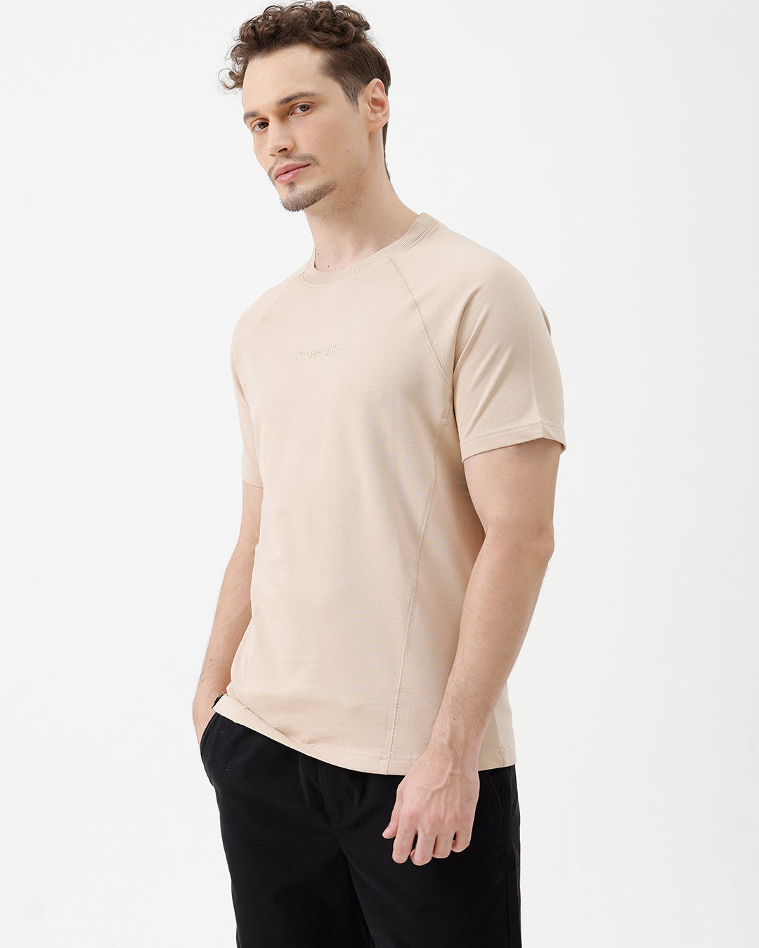 A man wearing a light beige short-sleeve t-shirt with a subtle tonal logo on the chest, paired with black pants, standing against a plain white studio background.