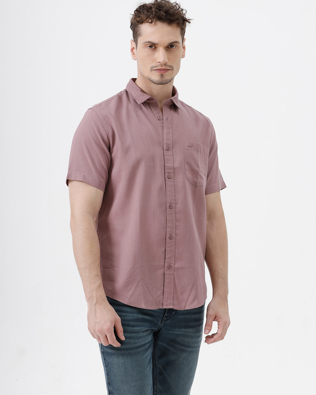 A man wearing a short-sleeve button-up shirt in dusty rose pink with dark blue jeans, standing against a plain white studio background.