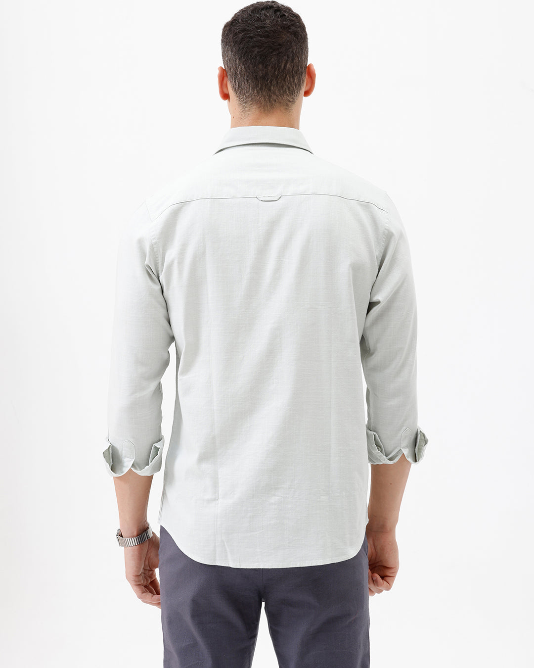 A man wearing a light sage green long-sleeve button-down shirt with rolled-up sleeves and dark gray trousers, standing against a plain white studio background.