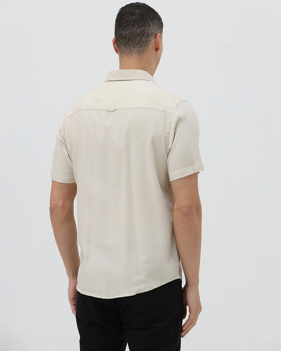 A man wearing a short-sleeve linen button-up shirt with a color-blocked design in beige and cream, paired with black pants, against a plain white studio background.