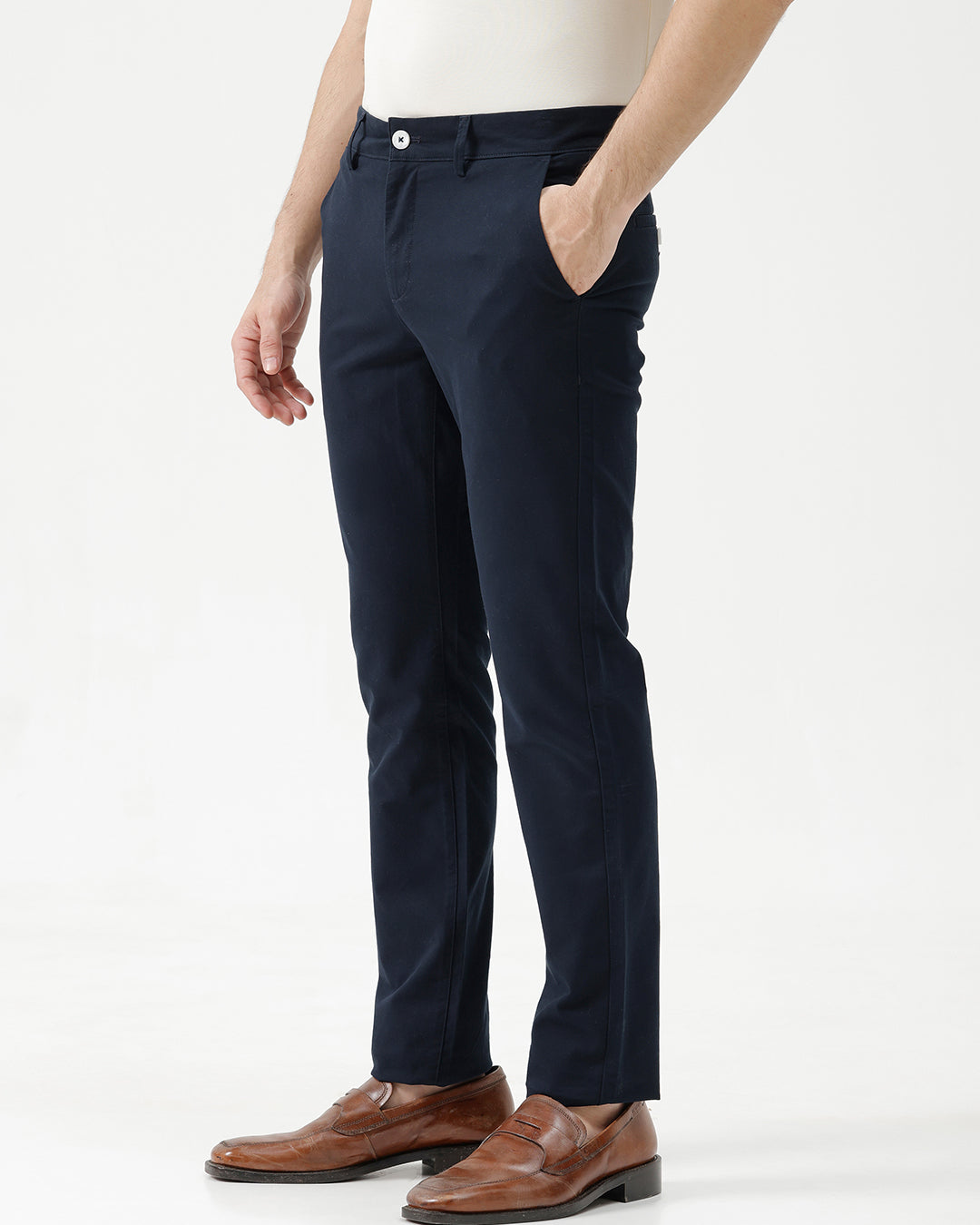 A man wearing dark navy blue slim-fit trousers, a cream-colored shirt, and brown leather loafers, standing against a plain white studio background.