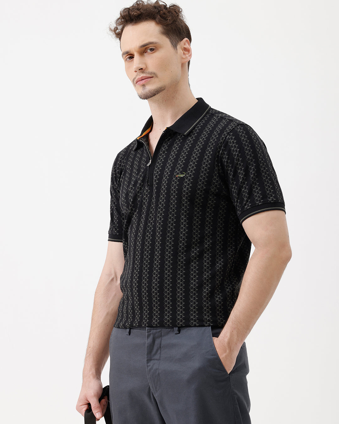 A man wearing a black short-sleeve polo shirt with a subtle geometric pattern, paired with dark gray trousers, standing against a plain white studio background.