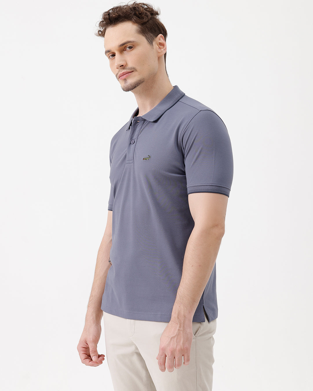 A man wearing a slate blue polo shirt and beige trousers, standing against a plain white background.