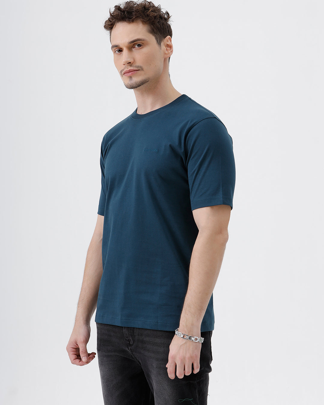 A man wearing a dark navy blue crew-neck t-shirt and black jeans, standing against a light gray studio background.
