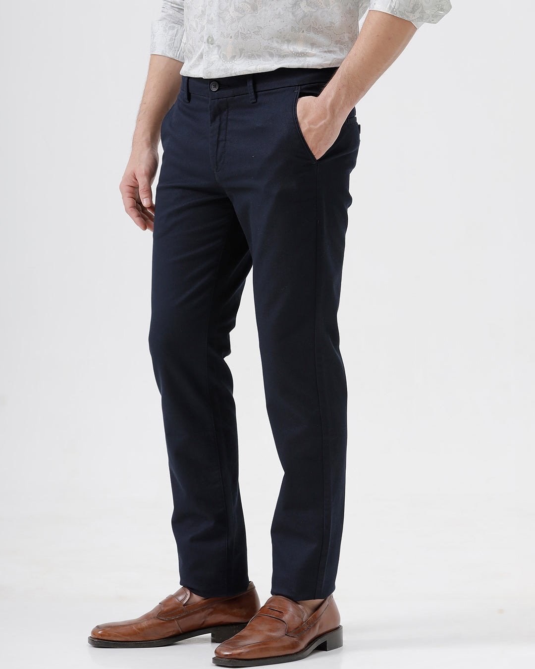A man wearing dark navy blue straight-leg trousers, a light-colored patterned shirt, and brown leather loafers, standing against a plain white studio background.