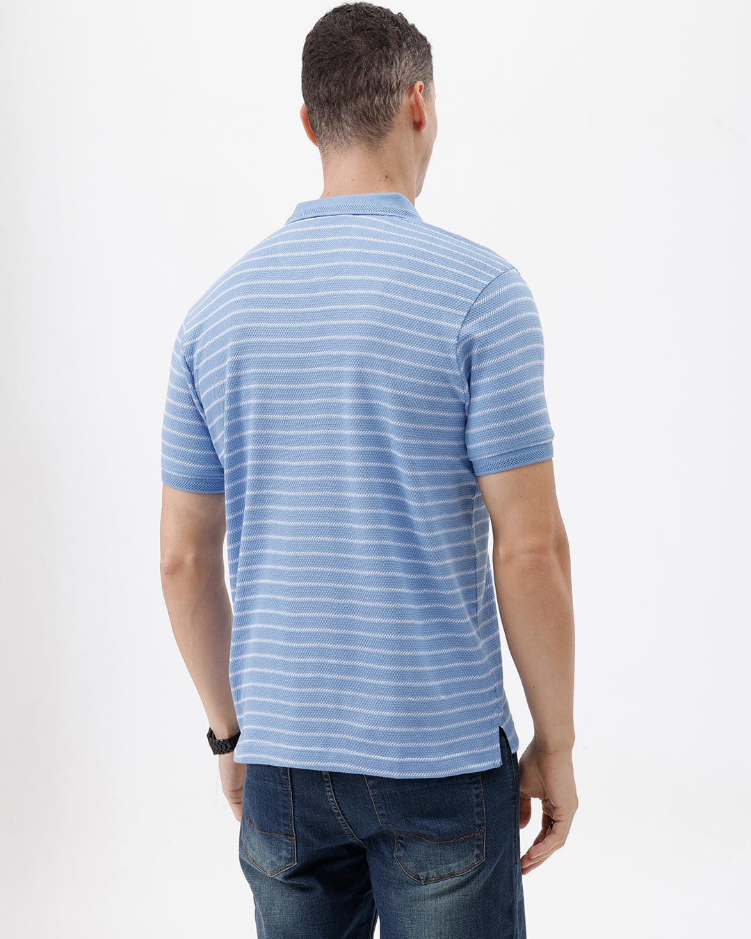 A man wearing a light blue and white striped short-sleeve polo shirt with dark blue jeans, standing against a plain light gray background.
