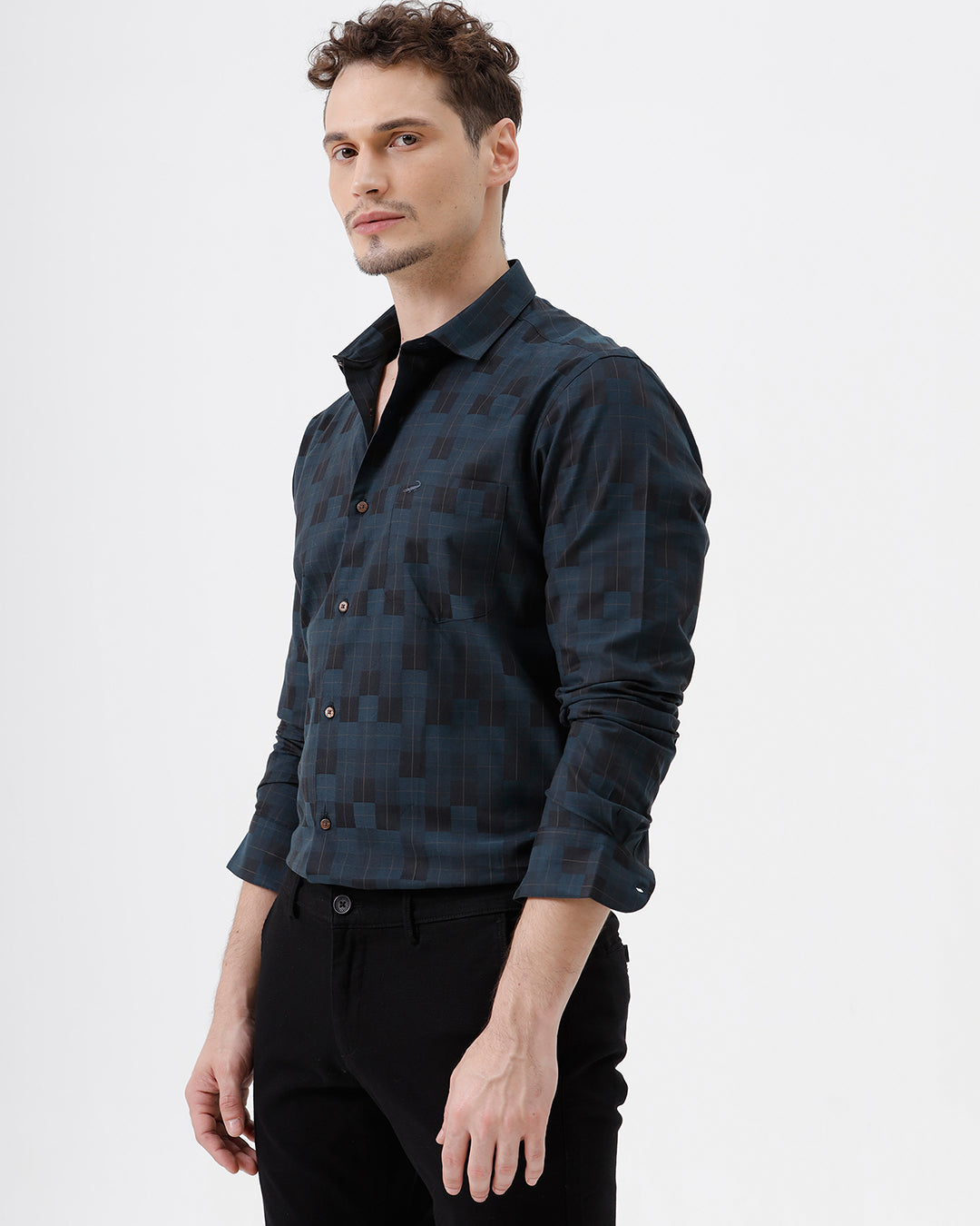 A man wearing a dark navy blue patterned long-sleeve button-up shirt and black trousers, standing against a plain white studio background.