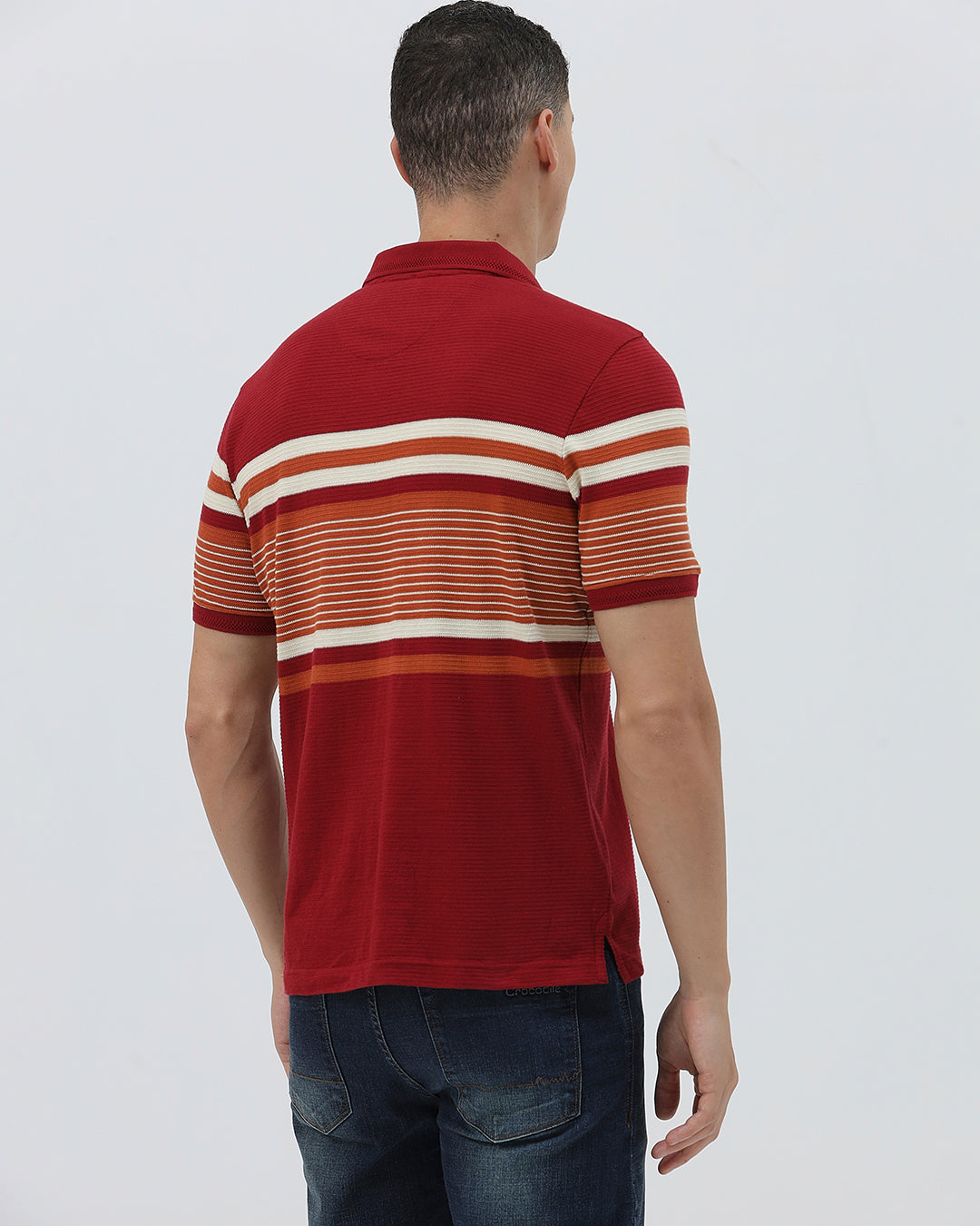 A man wearing a red short-sleeve polo shirt with horizontal cream and rust-orange stripes across the chest and sleeves, paired with dark blue jeans and black sunglasses, against a light gray studio background.