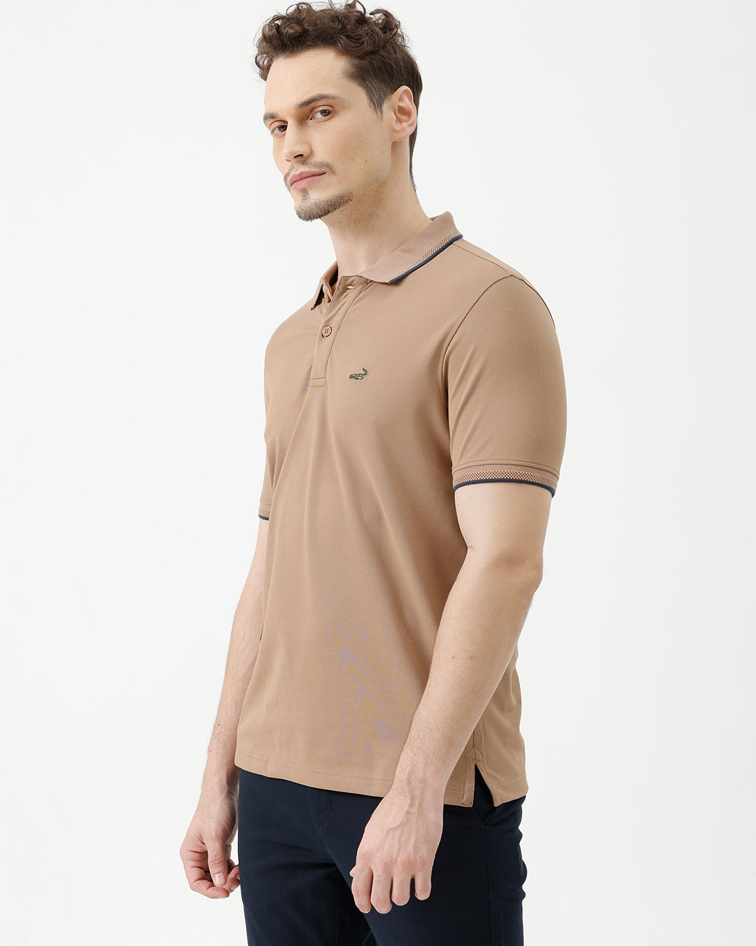 A man wearing a tan short-sleeve polo shirt with dark blue piping on the collar and sleeve cuffs, paired with dark navy pants, against a plain light gray background.
