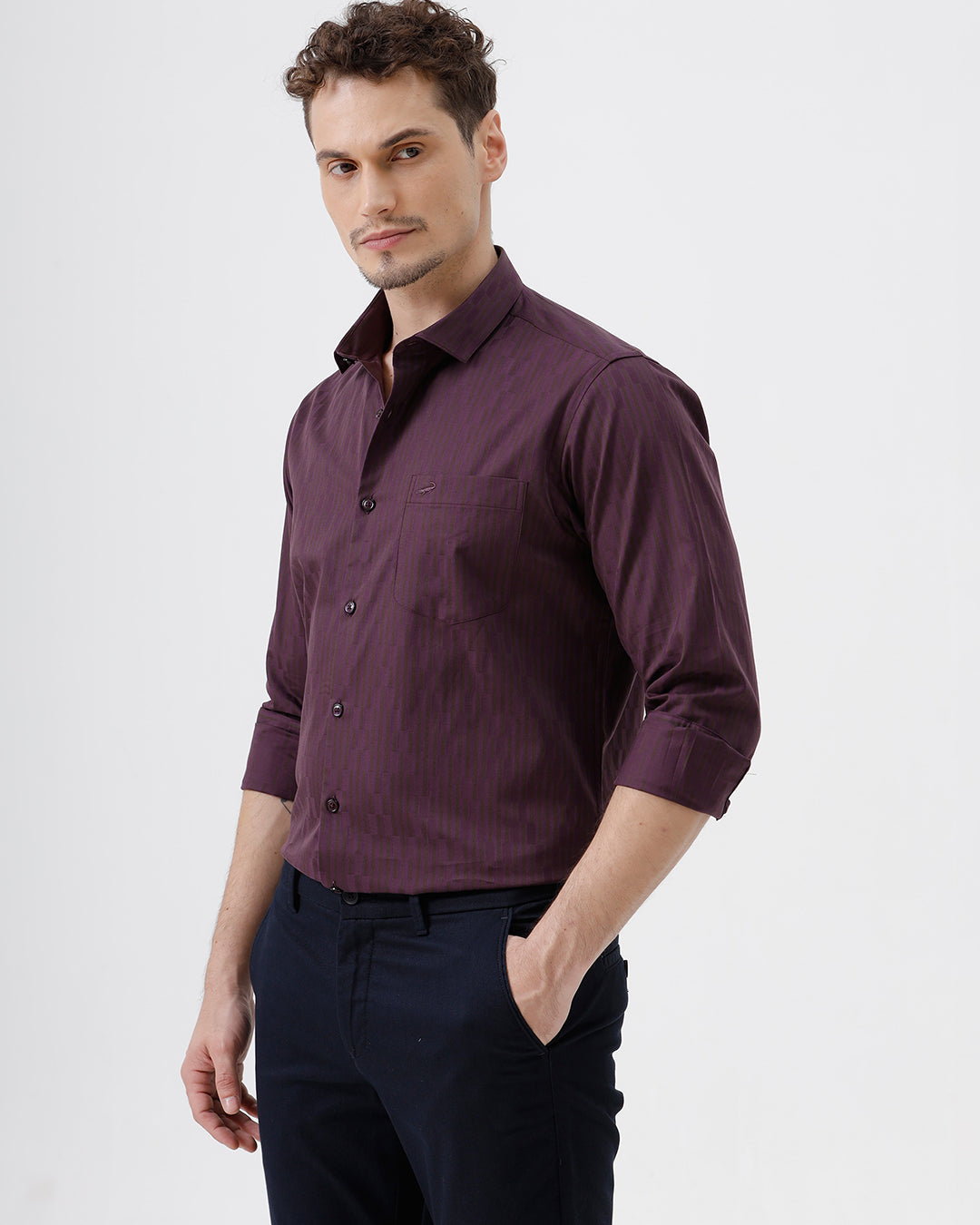 A man wearing a dark purple long-sleeve button-down shirt with subtle vertical stripes and dark navy trousers, standing against a light gray background.