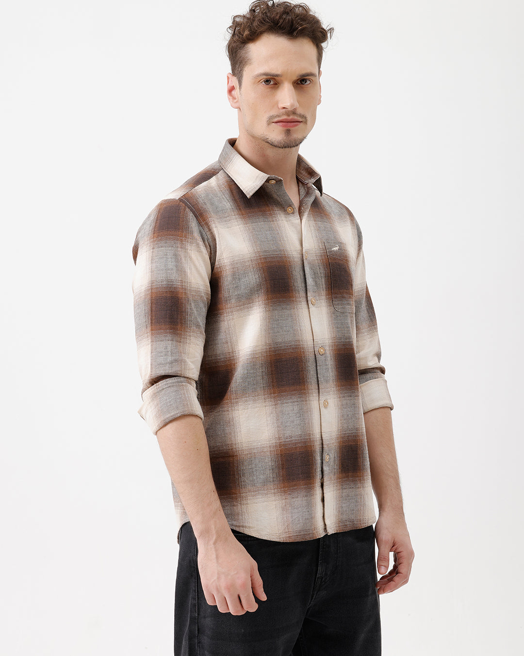A man wearing a long-sleeve button-up plaid shirt in brown, beige, and gray tones, paired with dark jeans, standing against a plain white studio background.