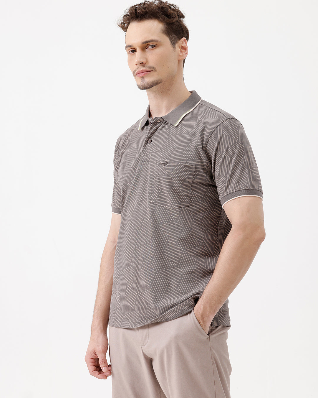 A man wearing a short-sleeve polo shirt with a gray and white geometric pattern, paired with beige trousers, standing against a plain white studio background.