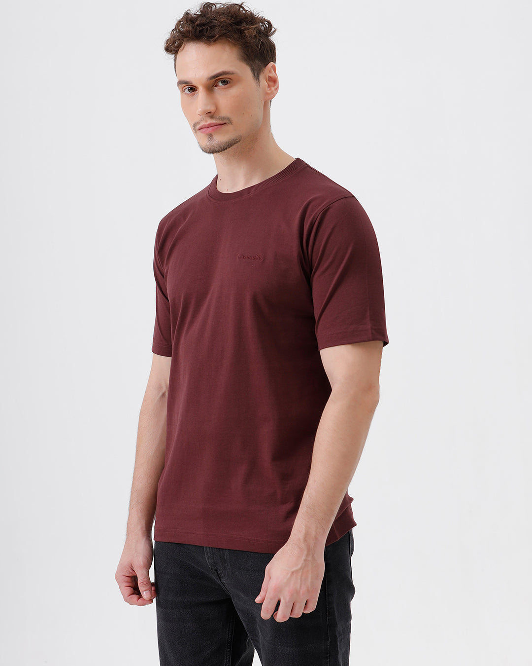 A man wearing a short-sleeve crewneck t-shirt in maroon, paired with dark jeans, standing against a plain light gray studio background.