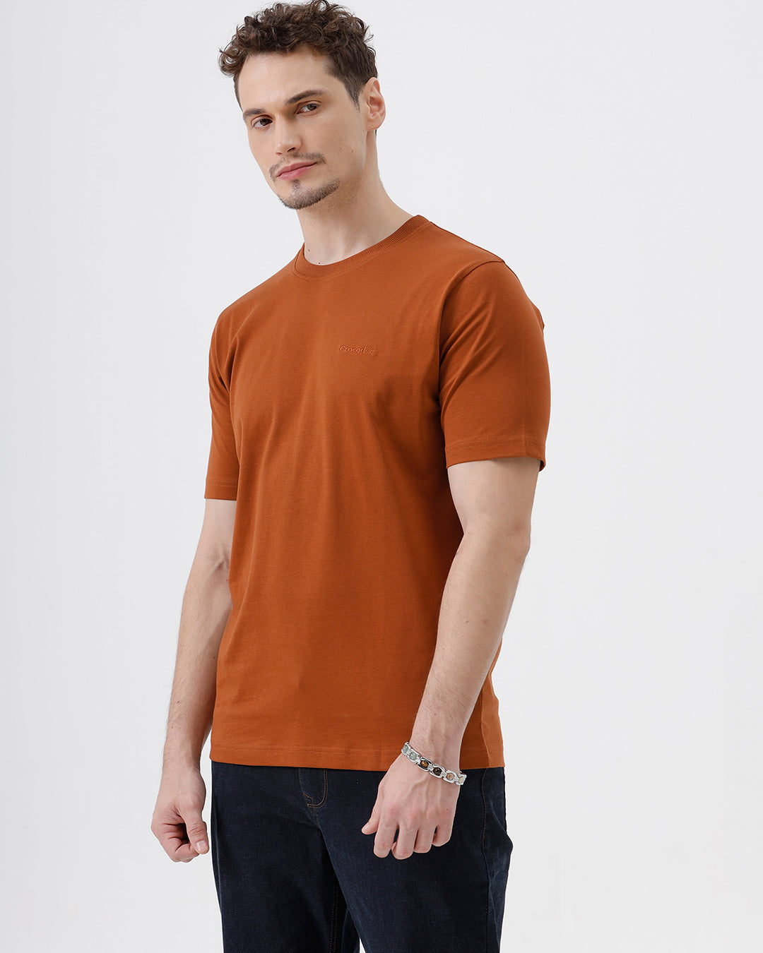 A man wearing a rust-orange crew-neck t-shirt and dark blue jeans, standing against a light gray studio background.