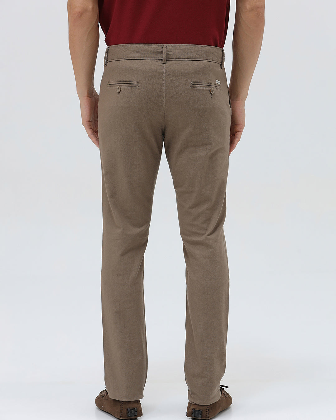 A person wearing slim-fit brown trousers, paired with a dark red shirt and brown suede moccasin shoes, shown from the waist down against a plain white studio background.