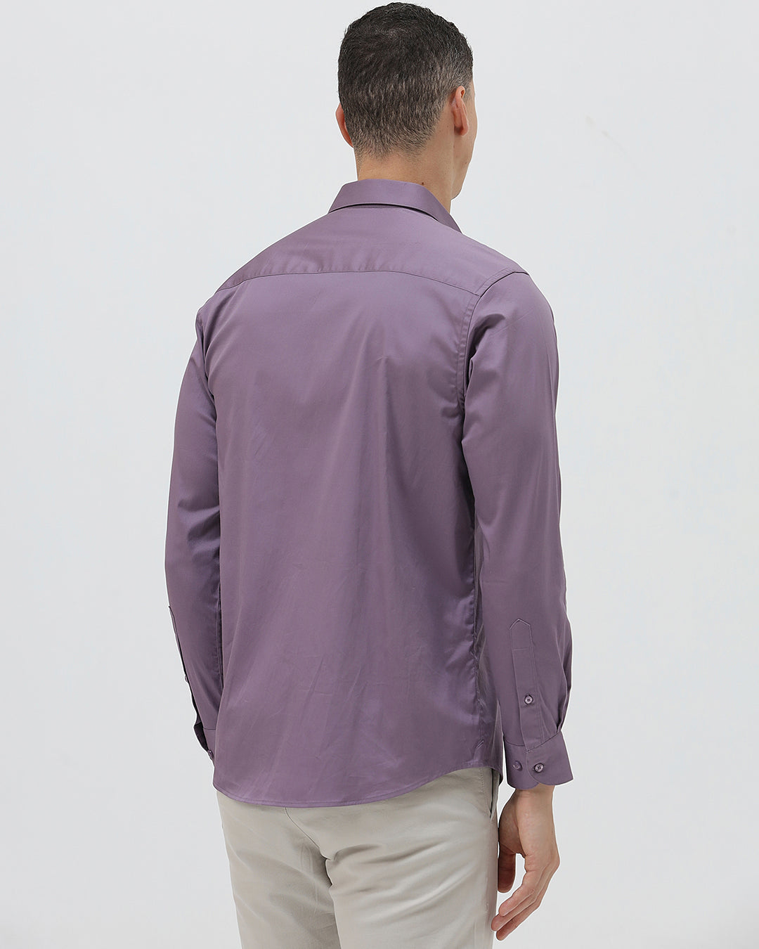 A man wearing a purple long-sleeve button-down shirt with rolled-up sleeves and light beige trousers, standing against a plain light gray background.