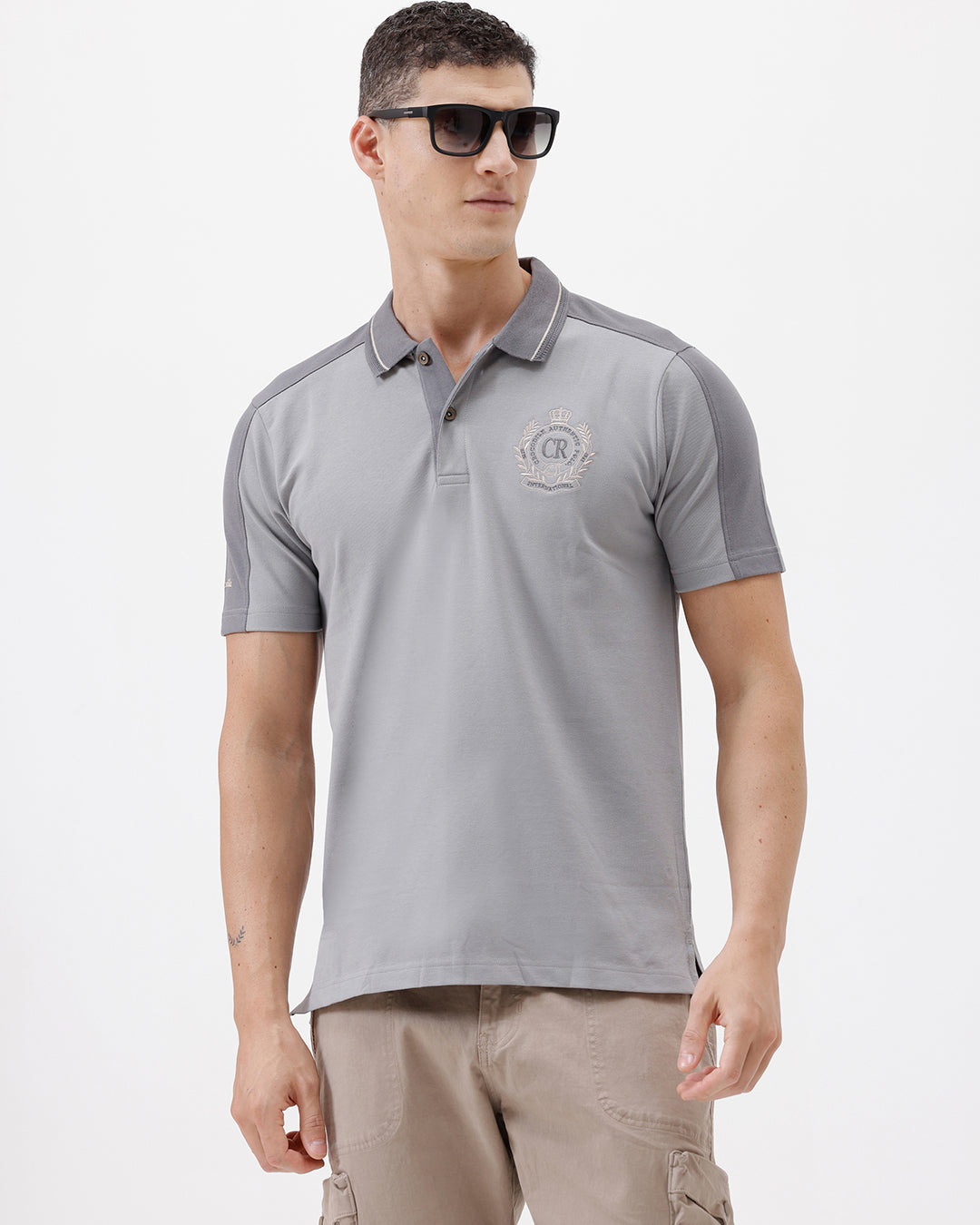 A man wearing a light gray short-sleeve polo shirt with darker gray shoulder panels and a small crest logo on the left chest, paired with khaki cargo pants, against a plain light gray studio background.