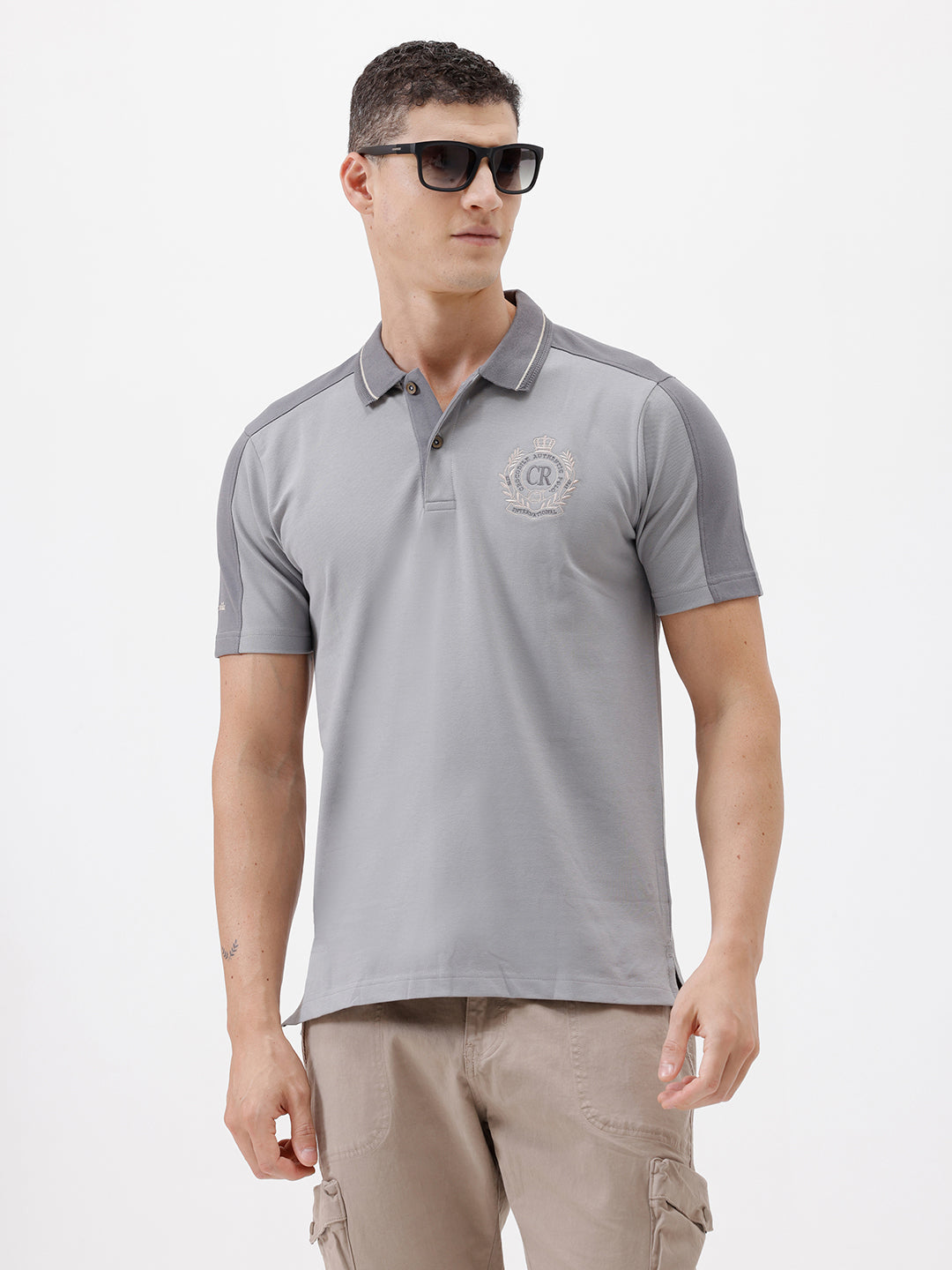 A man wearing a light gray short-sleeve polo shirt with darker gray shoulder panels and a small crest logo on the left chest, paired with khaki cargo pants, against a plain light gray studio background.