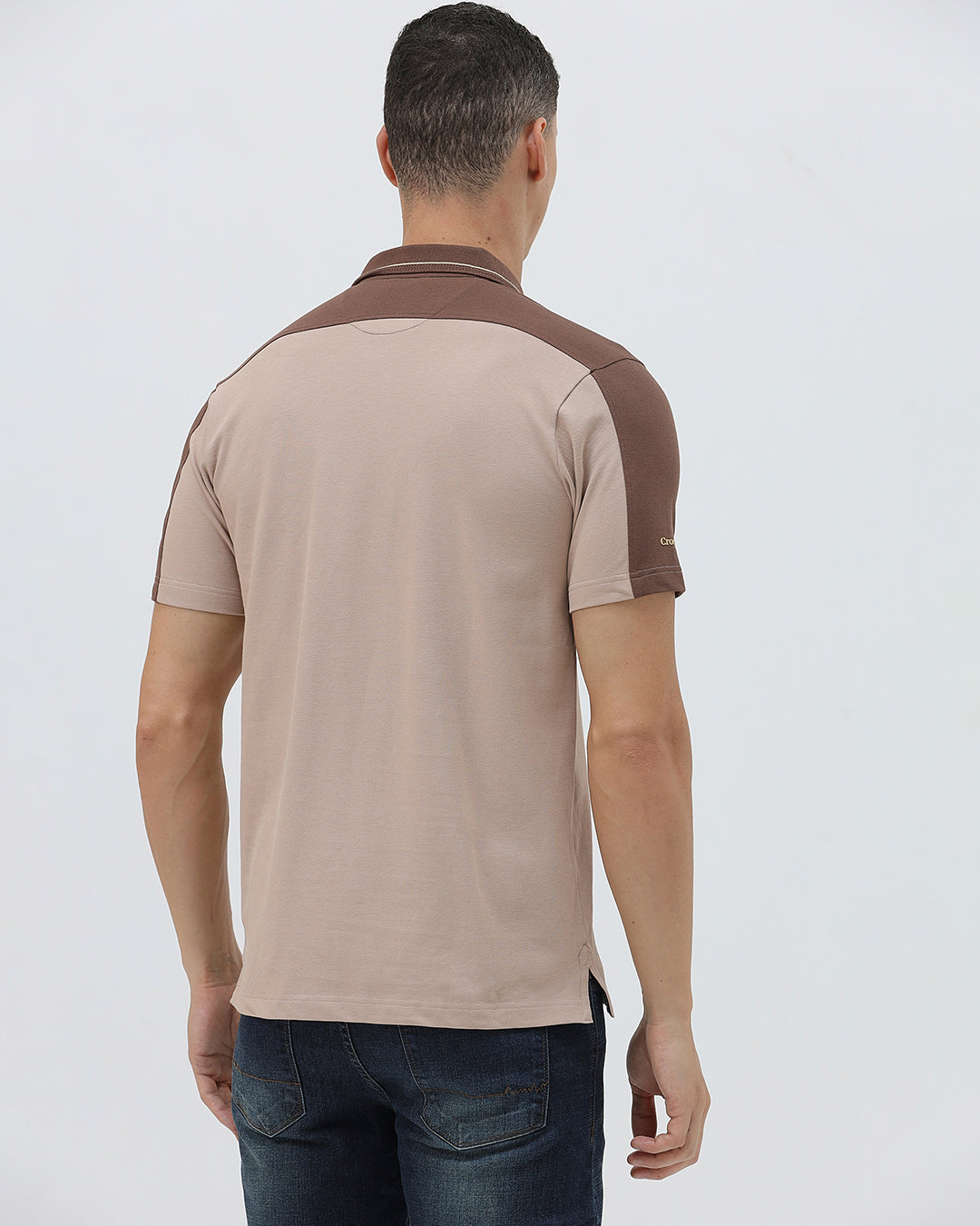 A man modeling a short-sleeve polo shirt in beige with brown shoulder panels and collar trim, paired with dark blue jeans, against a light gray studio background.