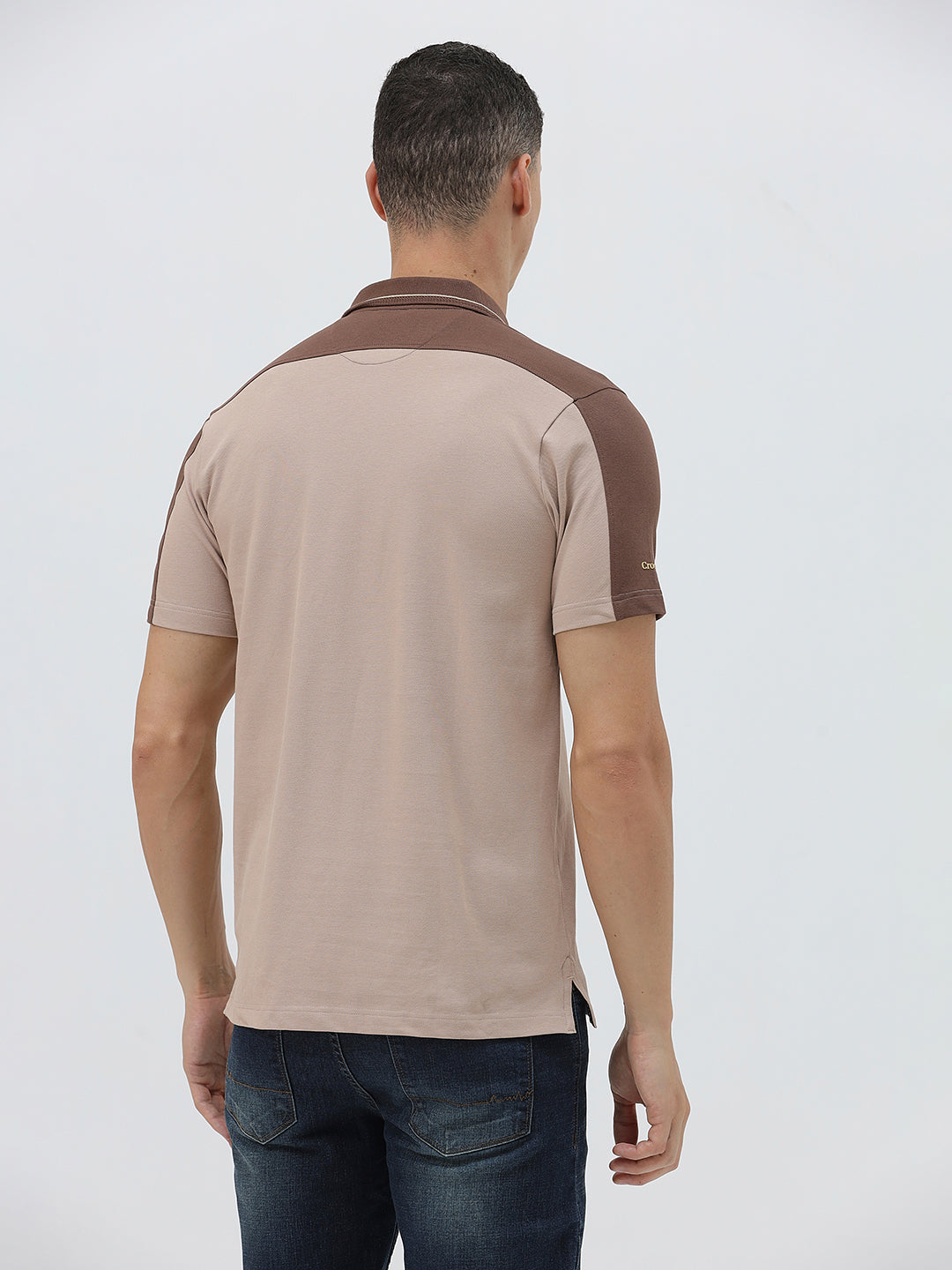 A man modeling a short-sleeve polo shirt in beige with brown shoulder panels and collar trim, paired with dark blue jeans, against a light gray studio background.