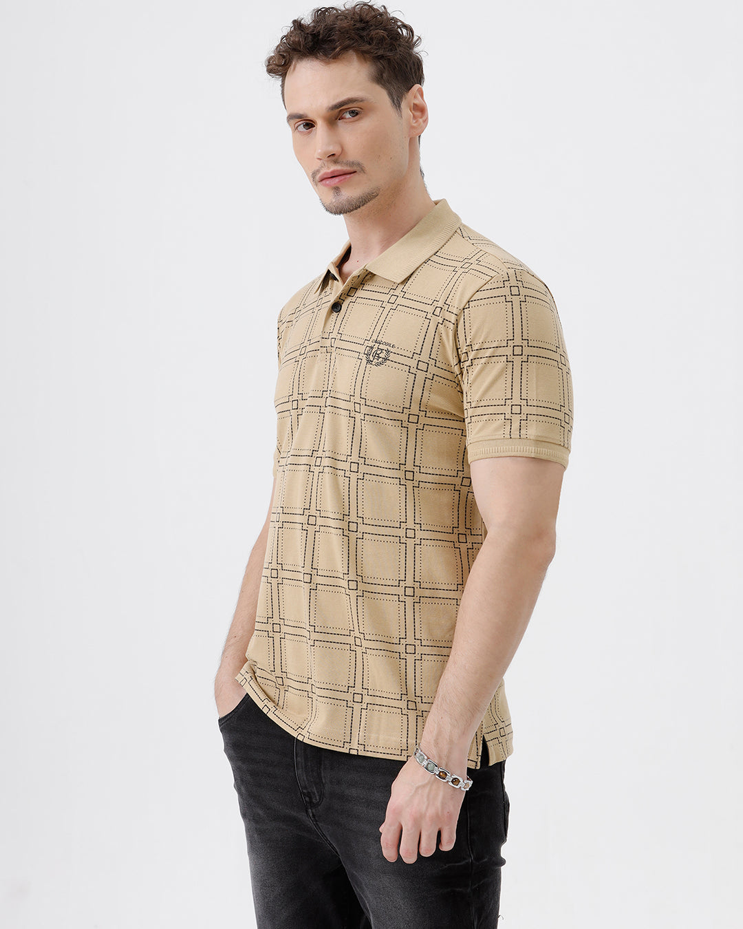 A man wearing a beige short-sleeve polo shirt with a brown grid pattern, paired with black jeans, standing against a plain white studio background.