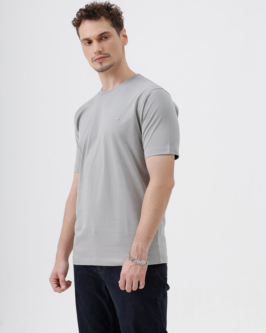 A man wearing a light gray short-sleeve crewneck t-shirt with a subtle tonal chest logo, paired with dark jeans, standing against a light gray studio background.