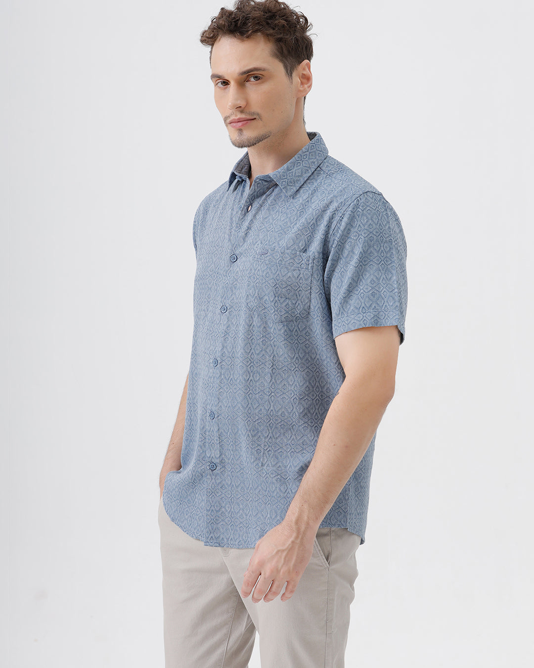 A man wearing a short-sleeve button-up shirt with a fine blue and white geometric pattern, paired with light beige trousers, standing against a plain light gray background.
