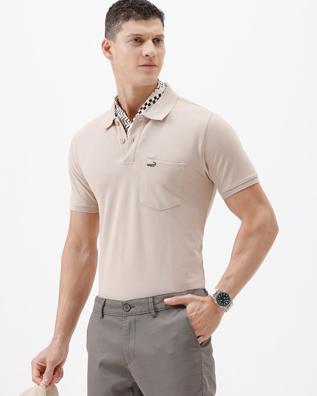 A man wearing a light beige short-sleeve polo shirt with a small crocodile logo on the chest pocket, paired with gray trousers, photographed against a plain white studio background.