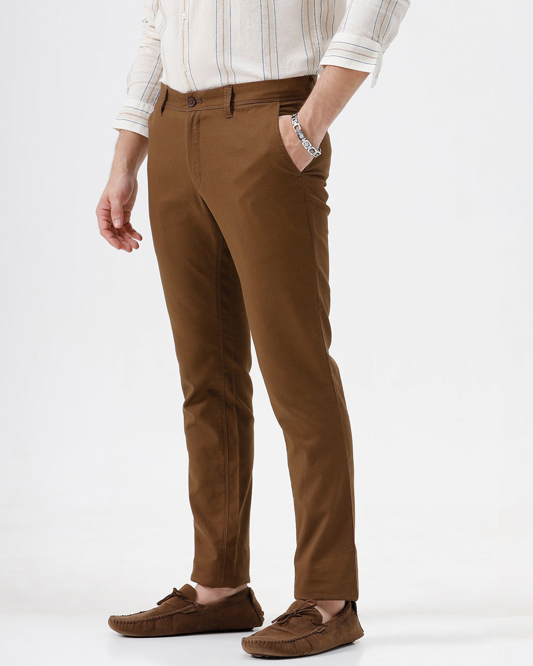 A man wearing brown slim-fit trousers, a cream and brown striped long-sleeve shirt, and brown suede loafers, standing against a plain white studio background.