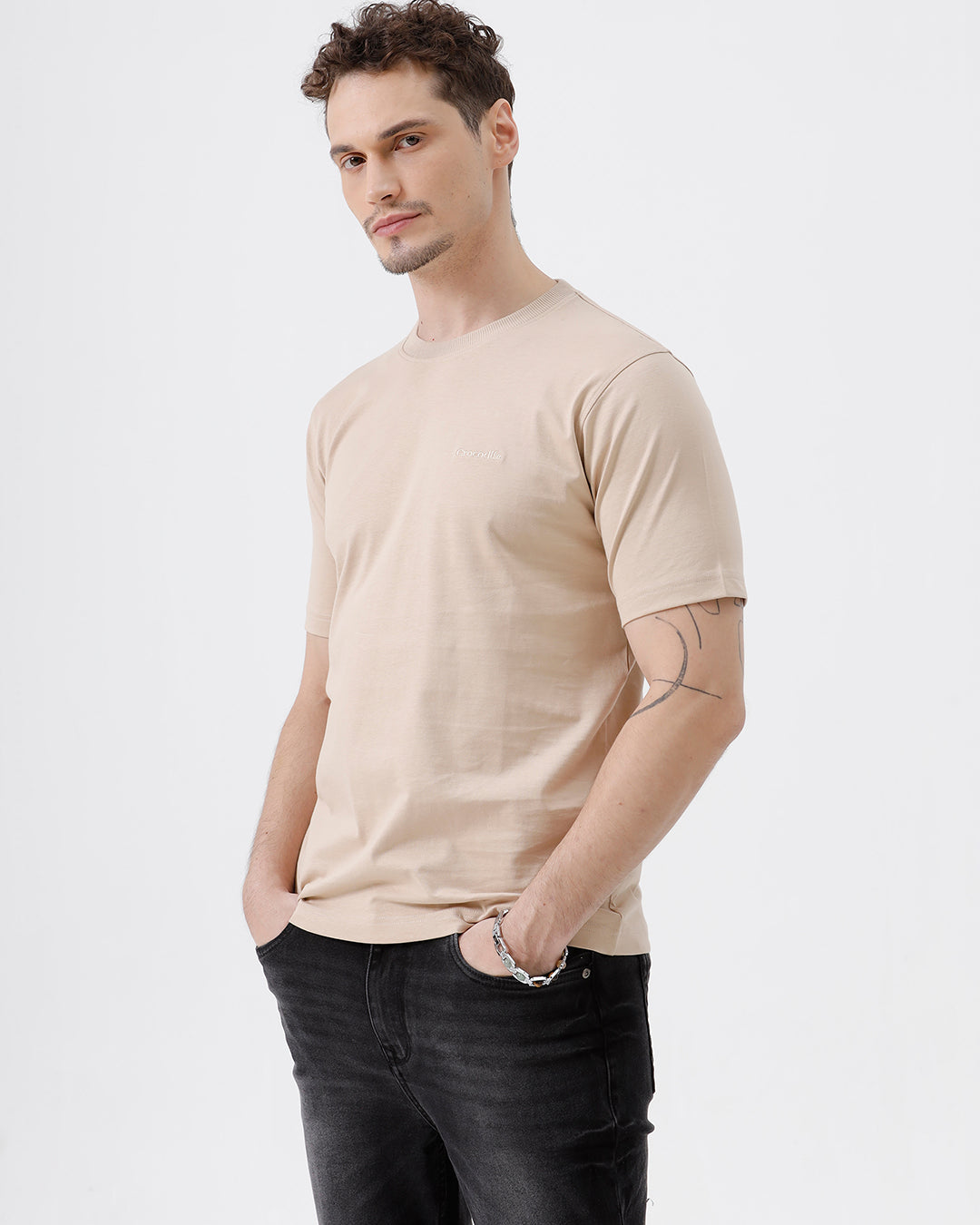 A man wearing a beige short-sleeve crewneck t-shirt with a subtle tonal logo on the left chest, paired with black jeans, standing against a light gray studio background.