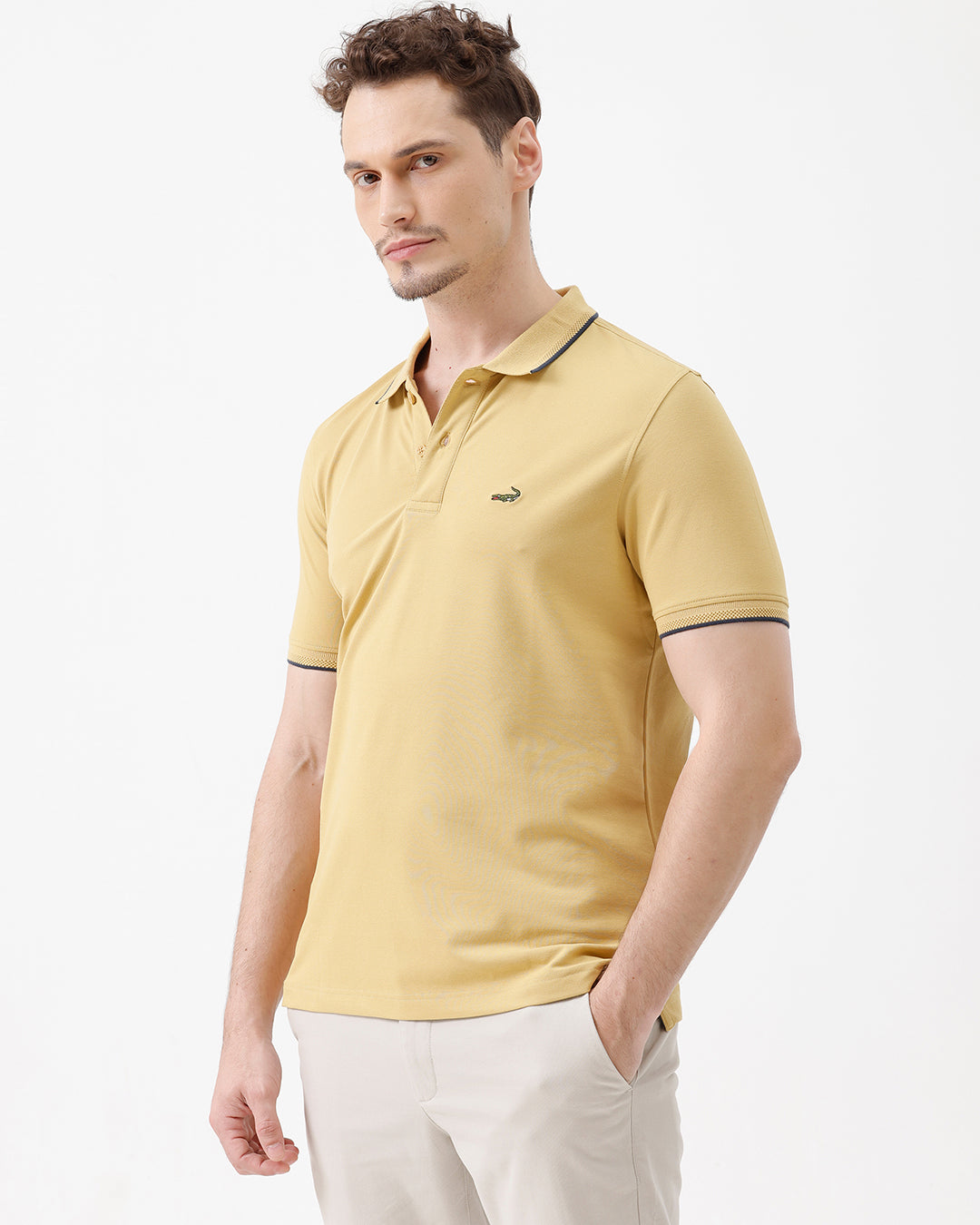 A man wearing a light yellow Lacoste polo shirt with dark piping on the collar and sleeve cuffs, paired with beige trousers, against a plain white studio background.