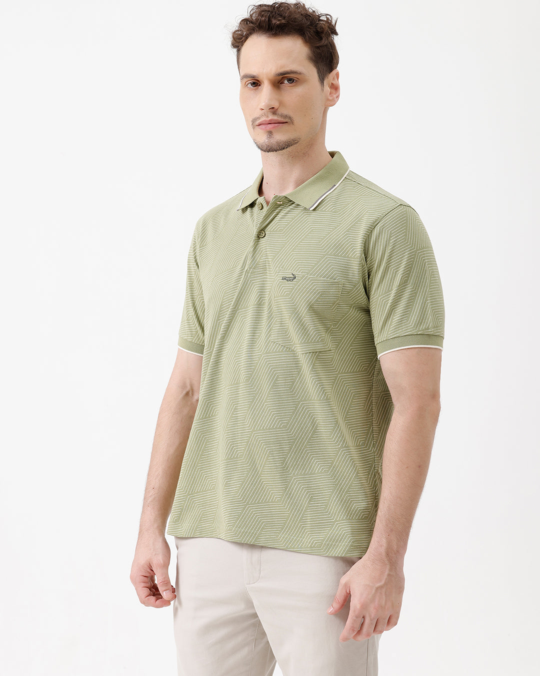A man wearing a light sage green short-sleeve polo shirt with a subtle geometric pattern, paired with beige trousers, standing against a plain white studio background.