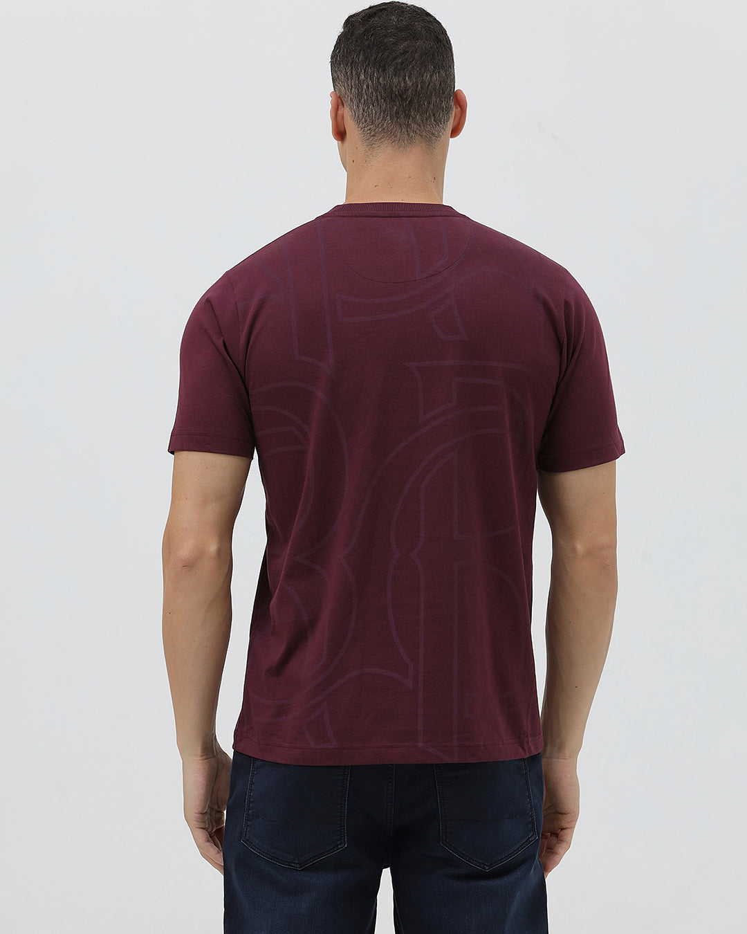 A man wearing a short-sleeve crewneck t-shirt in maroon with a subtle tonal graphic pattern, paired with dark blue jeans, against a light gray studio background.