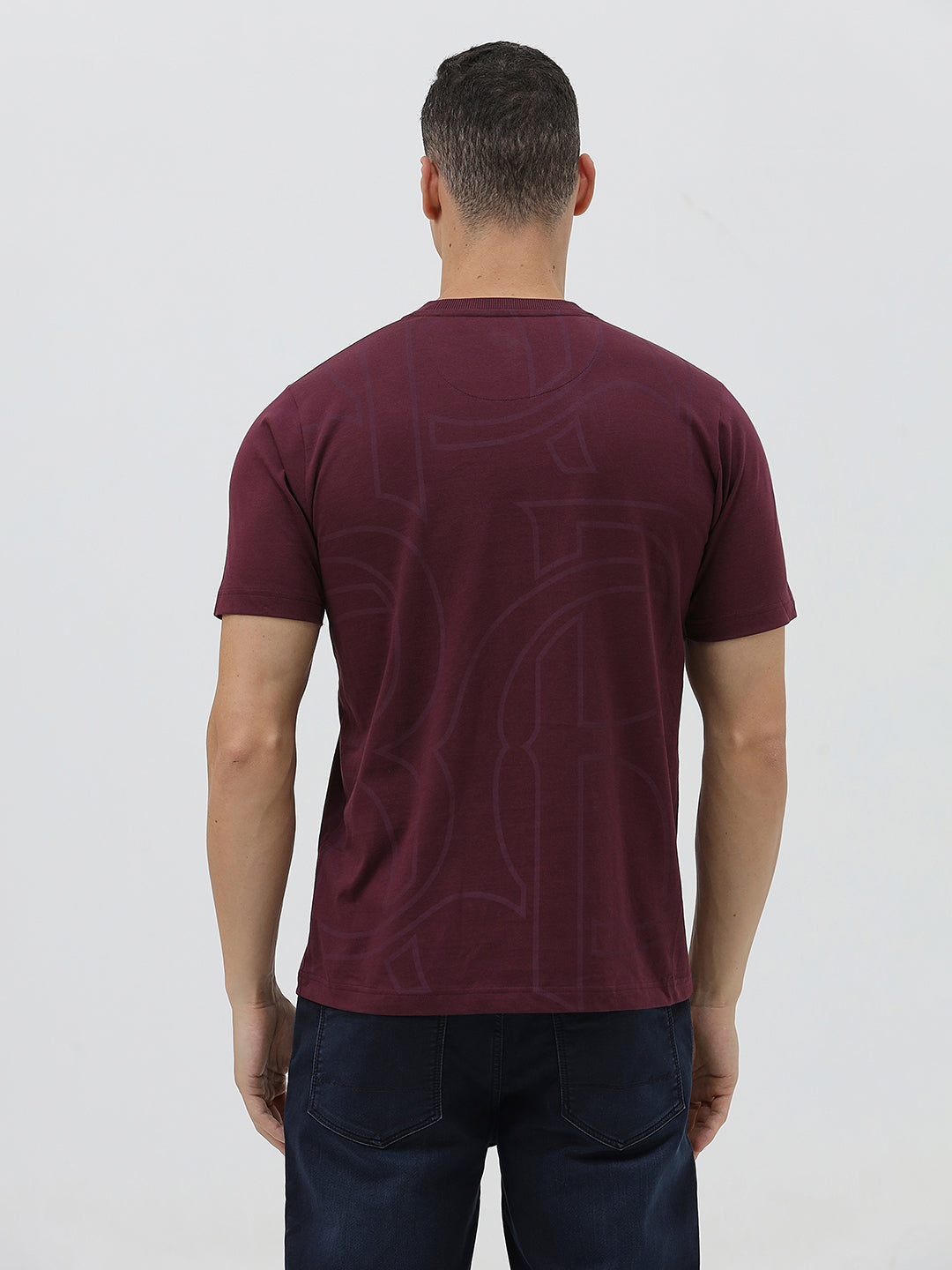 A man wearing a short-sleeve crewneck t-shirt in maroon with a subtle tonal graphic pattern, paired with dark blue jeans, against a light gray studio background.