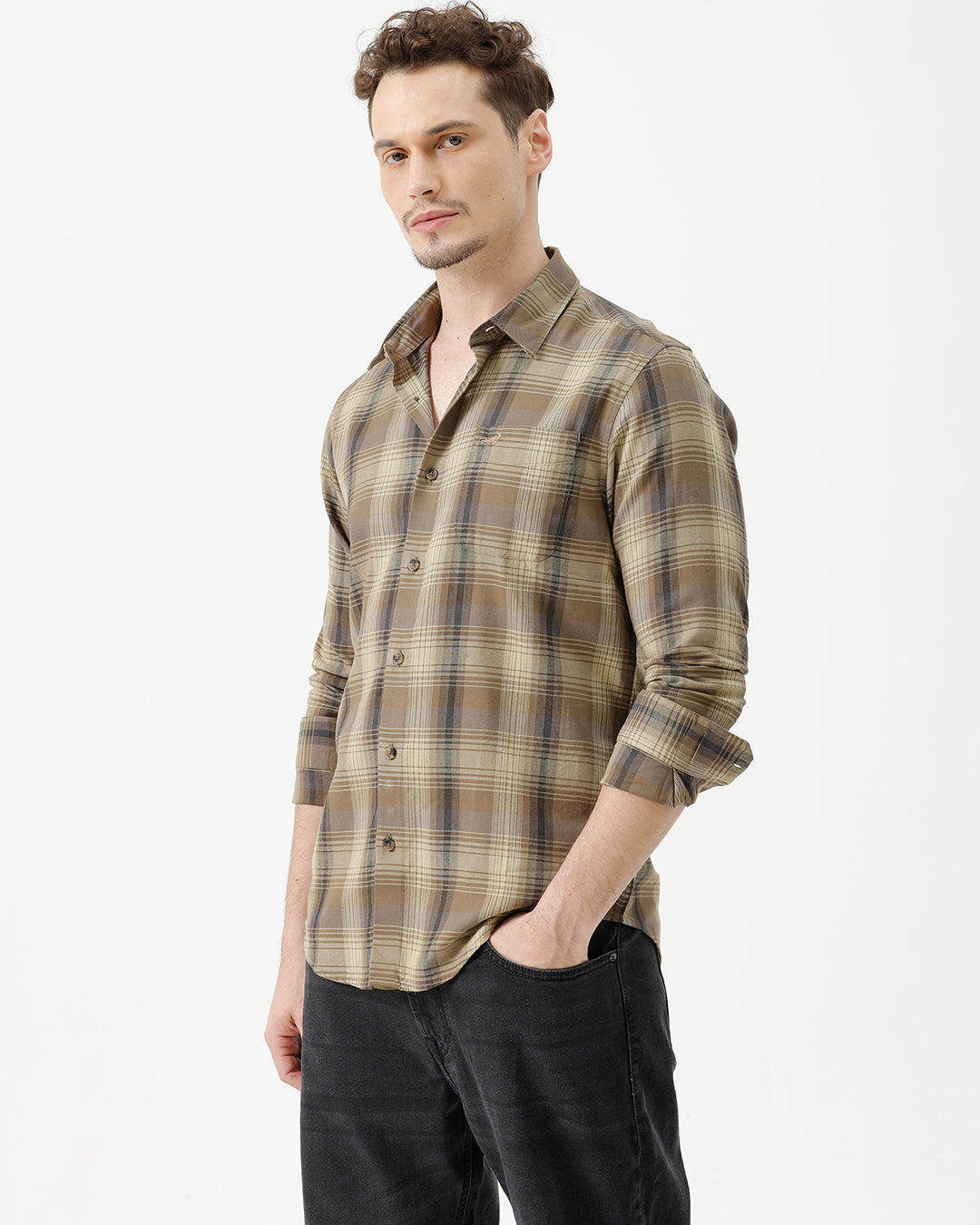 A man wearing a brown and beige plaid long-sleeve button-down shirt with dark sunglasses and black jeans, standing against a plain white studio background.