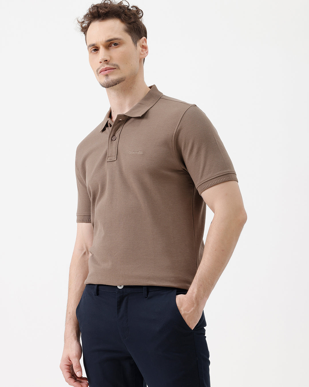 A man wearing a brown short-sleeve polo shirt with dark navy pants, standing against a plain light gray studio background.