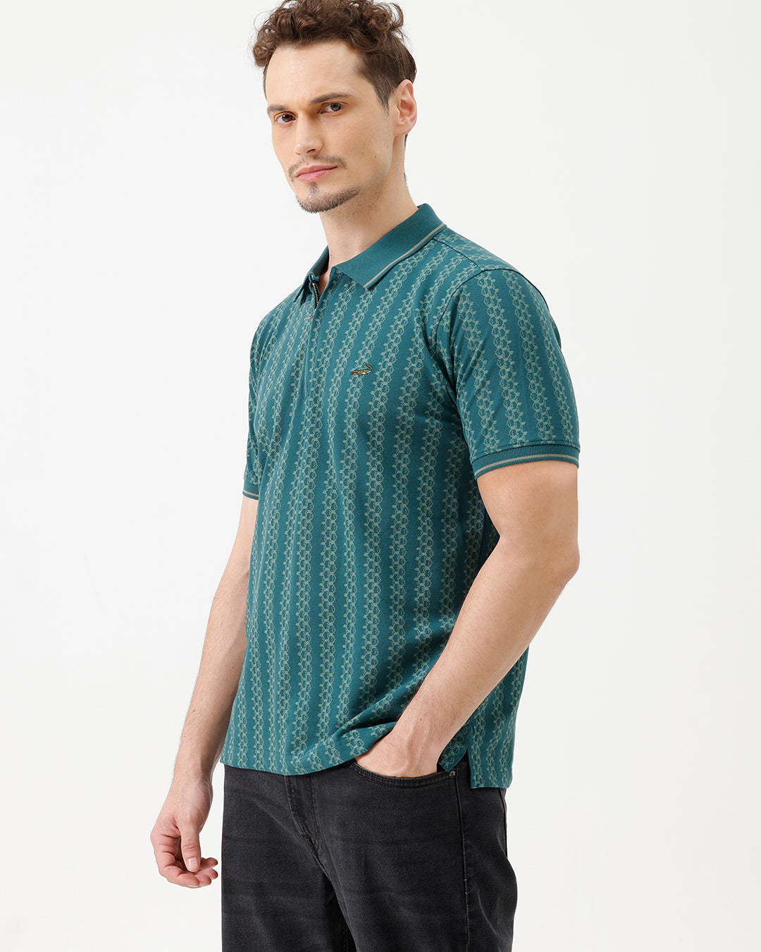 A man wearing a teal green short-sleeve polo shirt with a white geometric pattern, paired with black jeans, standing against a plain white studio background.