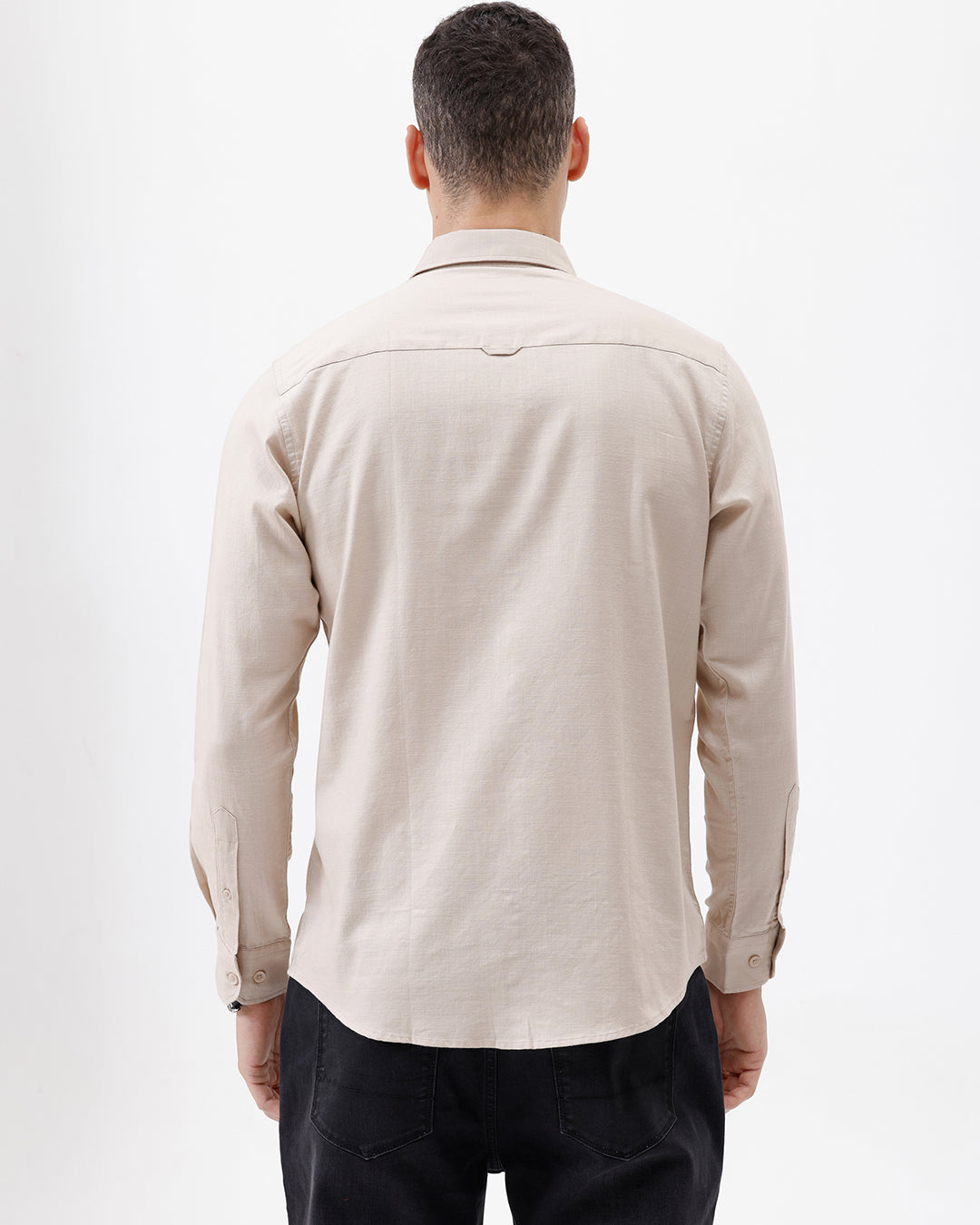 A man wearing a light beige long-sleeve button-up shirt with a chest pocket, paired with dark jeans, standing against a plain white studio background.