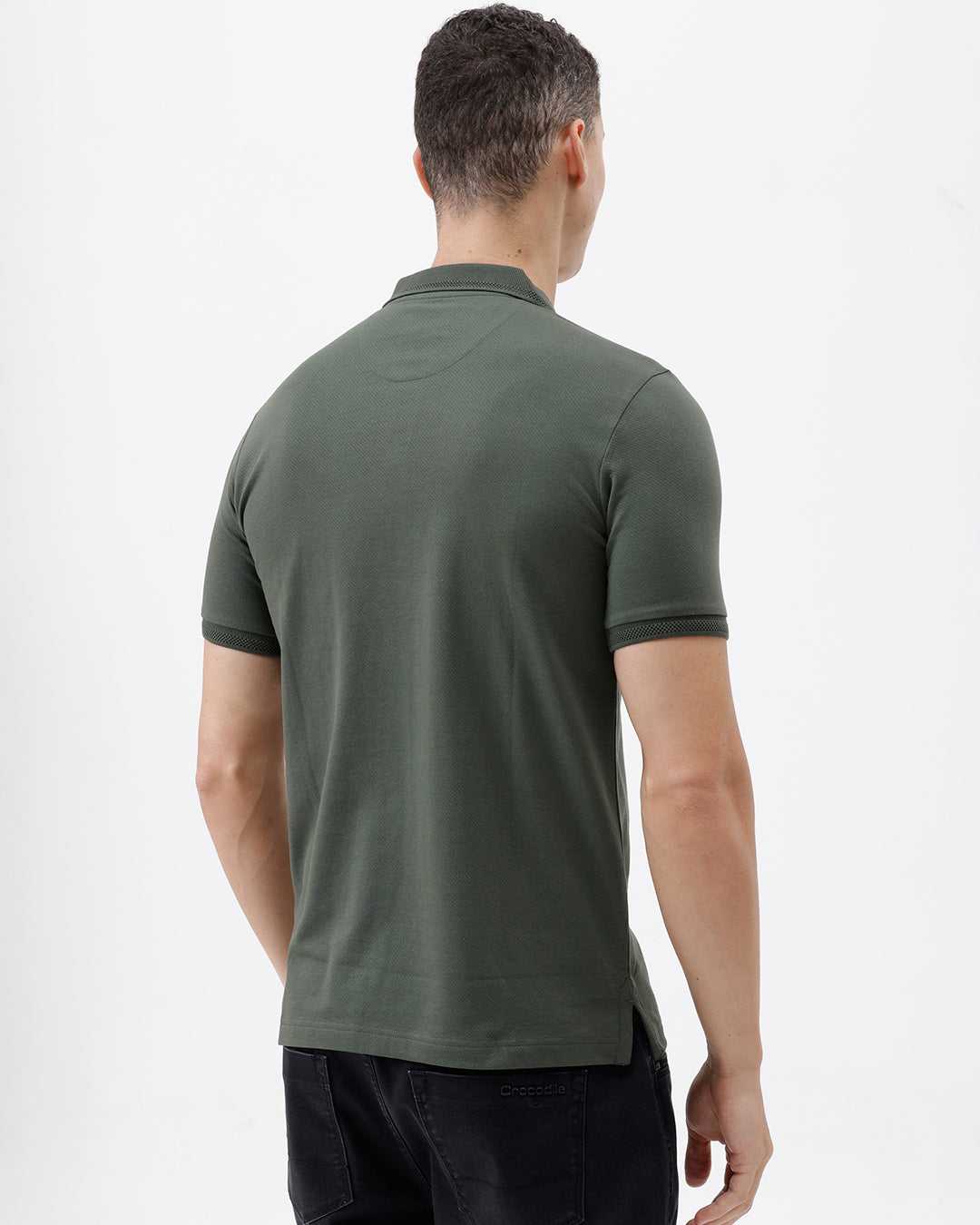A man wearing a dark olive green short-sleeve polo shirt with a chest pocket, paired with dark jeans, standing against a plain light gray background.