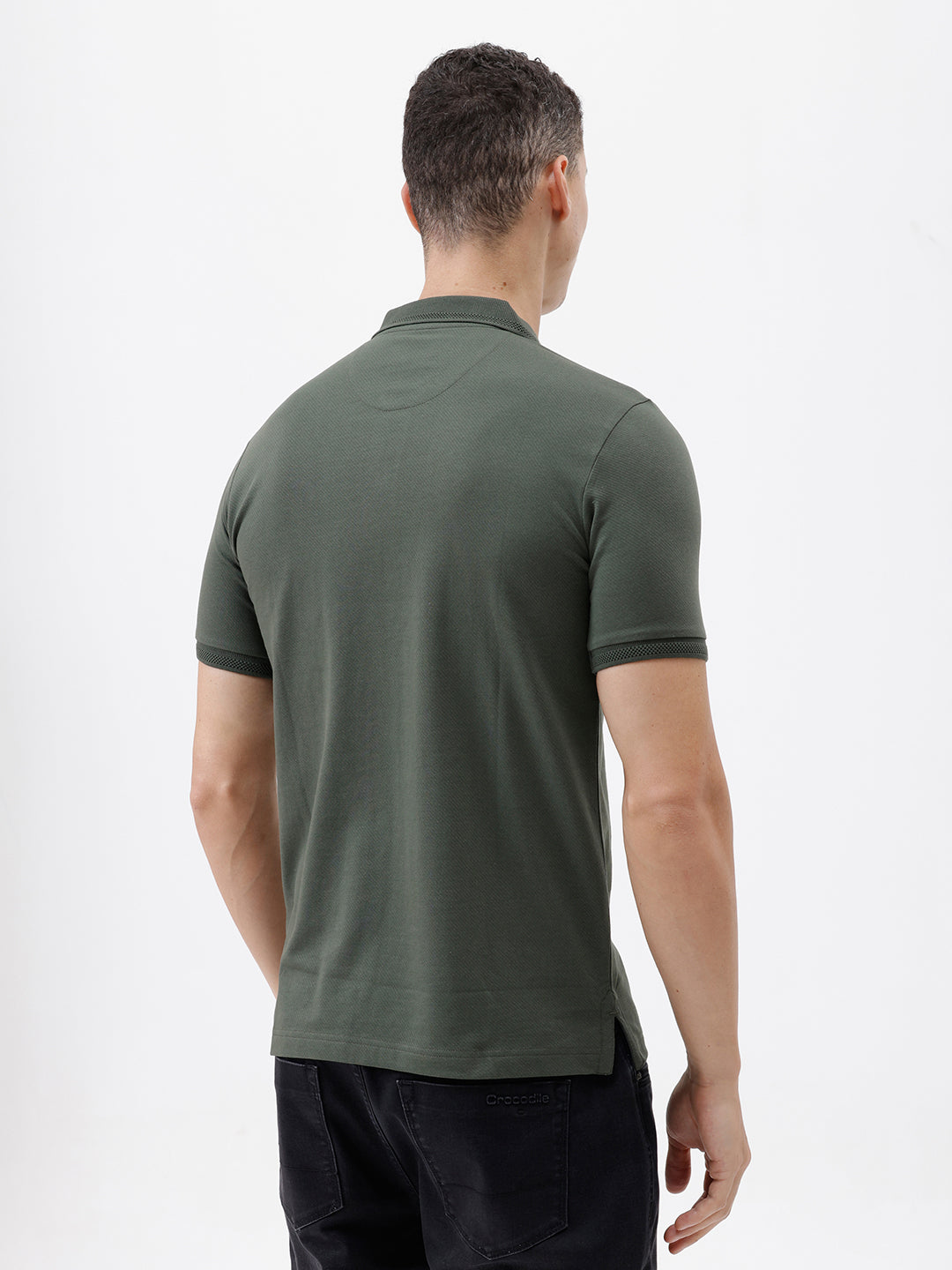 A man wearing a dark olive green short-sleeve polo shirt with a chest pocket, paired with dark jeans, standing against a plain light gray background.