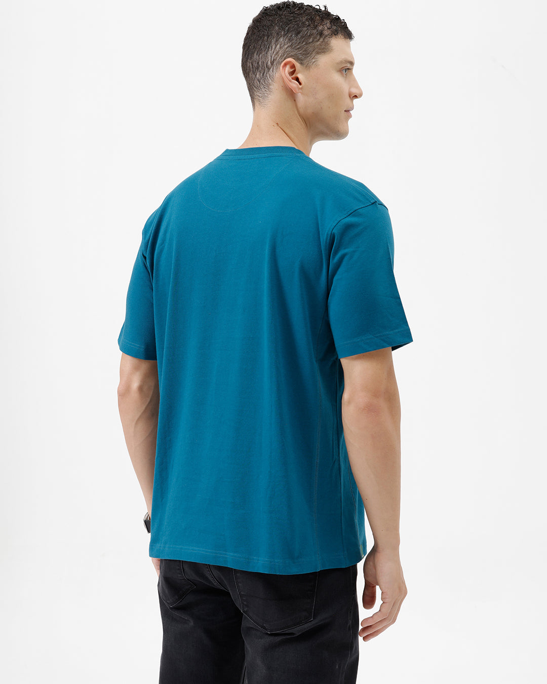 A man wearing a teal blue short-sleeve t-shirt with a subtle tonal 'GREATNESS' text graphic across the chest, paired with black pants, photographed against a clean white studio background.