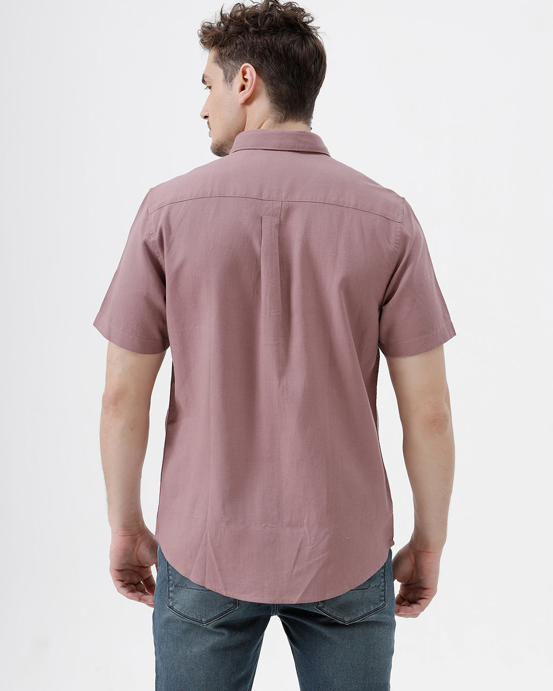 A man wearing a short-sleeve button-up shirt in dusty rose pink with dark blue jeans, standing against a plain white studio background.