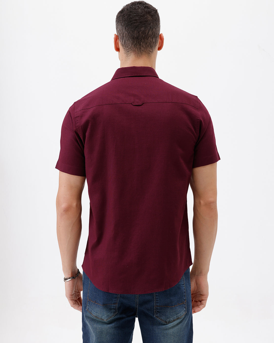 A man wearing a short-sleeve button-up shirt in maroon with a chest pocket, paired with blue jeans, standing against a plain white studio background.