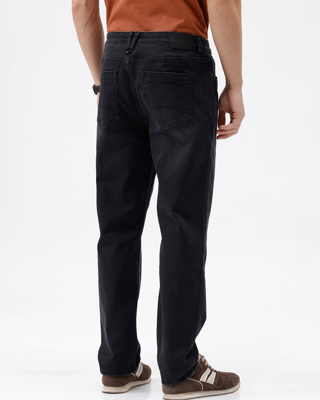 A person wearing black slim-fit jeans, paired with a rust-orange t-shirt and brown sneakers, shown from the waist down against a plain white studio background.