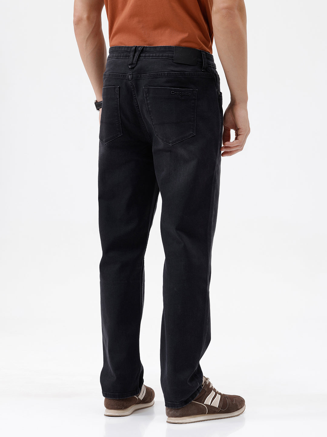 A person wearing black slim-fit jeans, paired with a rust-orange t-shirt and brown sneakers, shown from the waist down against a plain white studio background.