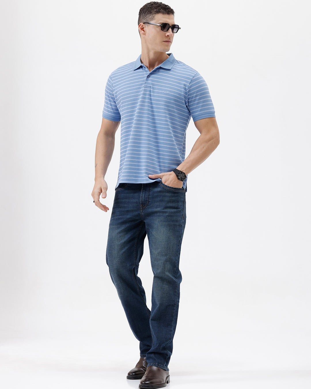 A man wearing a light blue and white striped short-sleeve polo shirt with dark blue jeans, standing against a plain light gray background.