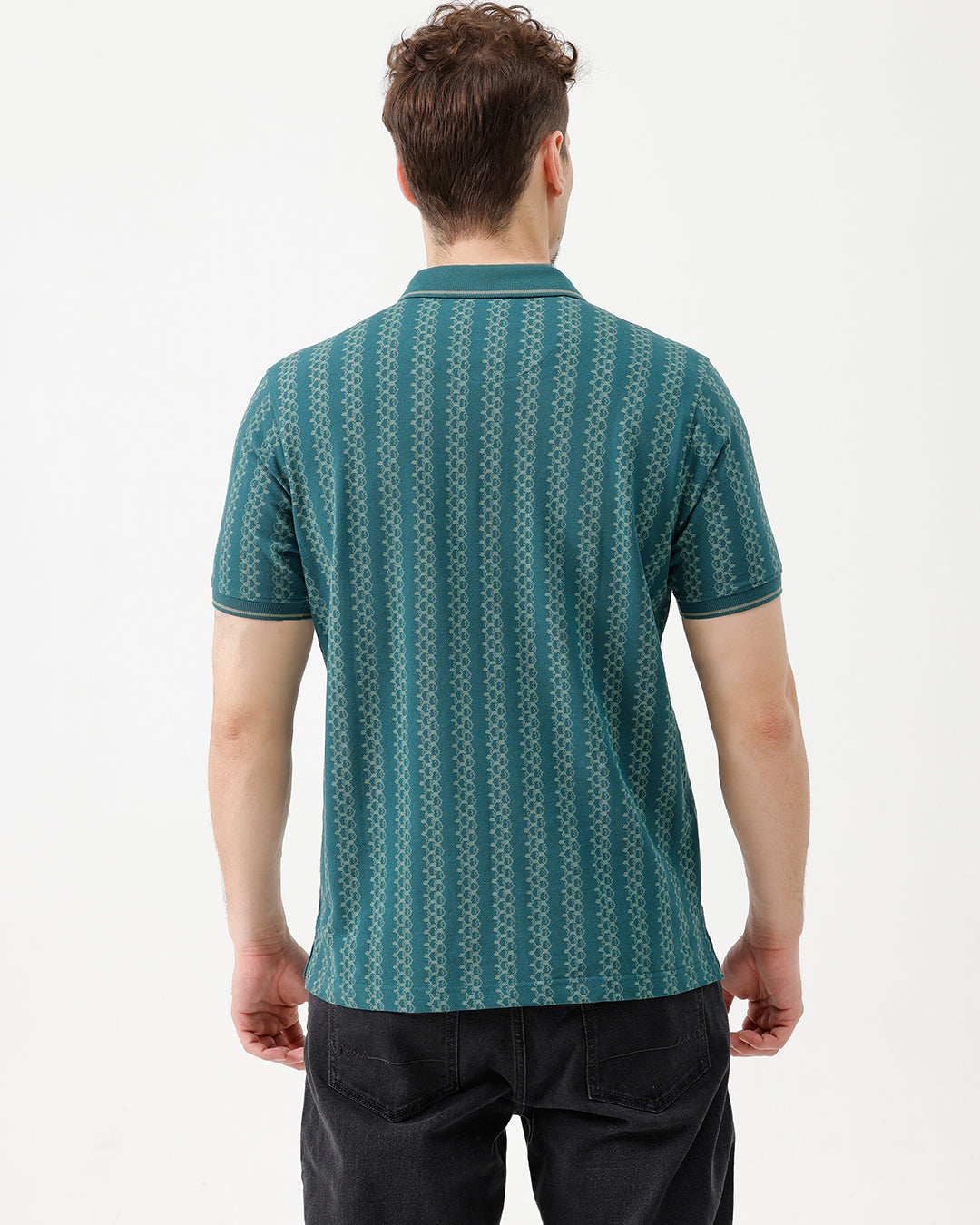 A man wearing a teal green short-sleeve polo shirt with a white geometric pattern, paired with black jeans, standing against a plain white studio background.